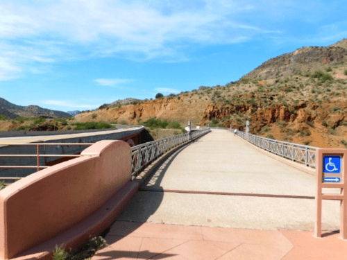 A paved bridge leads into a scenic landscape with hills and blue sky, featuring a wheelchair access sign.